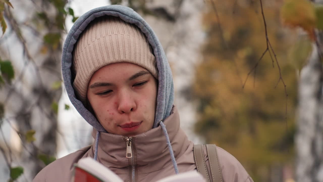Light skin lady in warm beige jacket and knit hat reading red book, licking lips thoughtfully while standing close to tree in autumn setting with softly blurred background and golden leaves