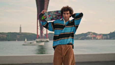 hombre joven posando patineta en el muelle de vista de agua. patinador mirando cámara vertical