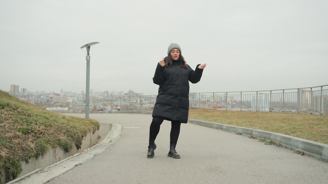 Siyah palto ve bere giymiş beyaz tenli bir kadın, tepe yolunda etkileyici bir dans sergiliyor; yavaş ritmik ayak hareketleri ve zarif kol jestleri, gri gökyüzü altındaki panoramik şehir manzarasıyla birleşerek iç gözlem ve dayanıklılık duyguları uyandırıyor.