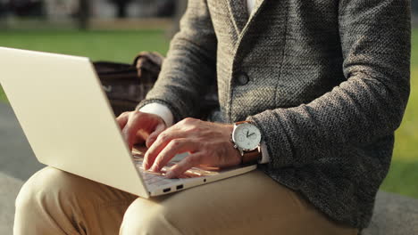 close up of a handsome businessman sitting on wall in the city park and working on his laptop computer