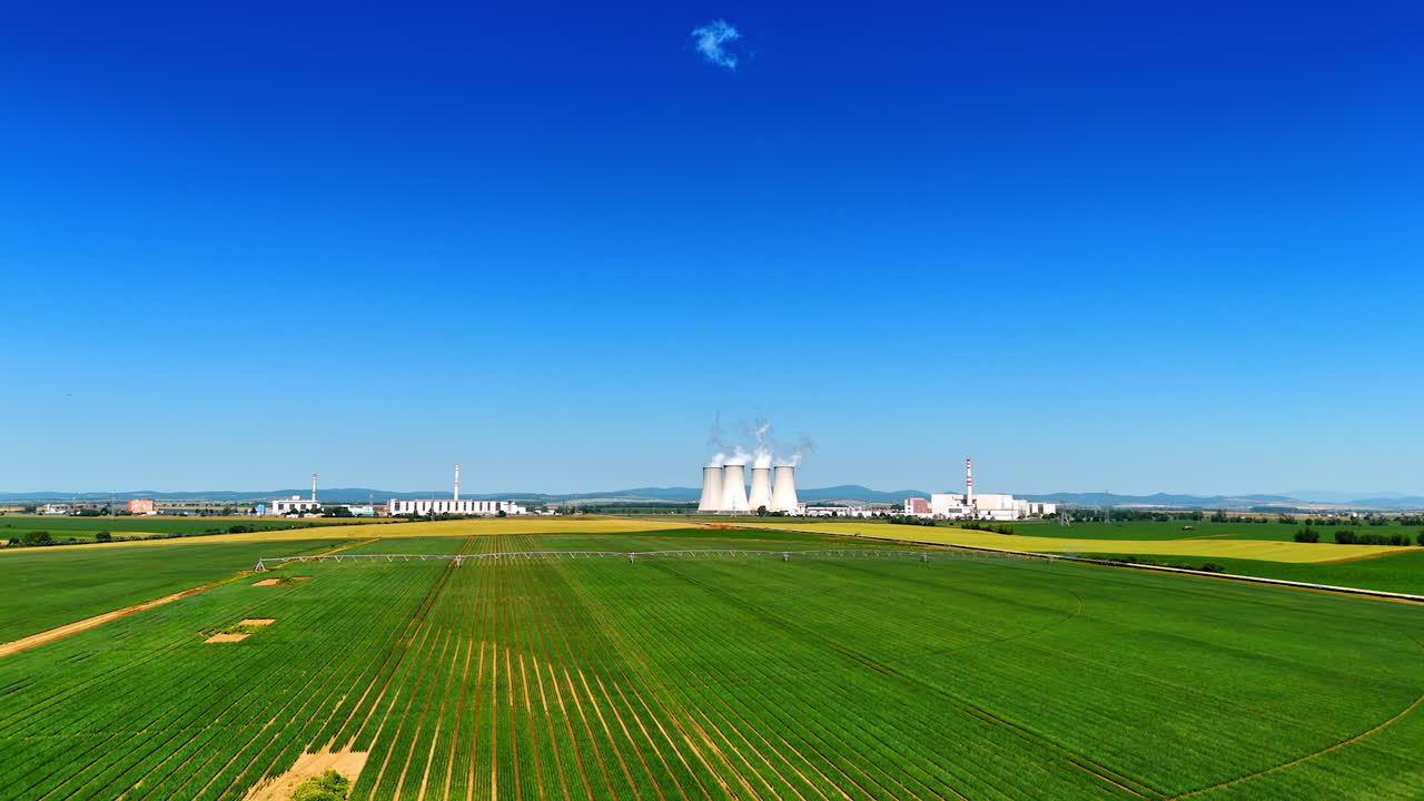 Green agricultural fields and contrasting blue clear sky. Premises and tubes of the plant at the horizon.