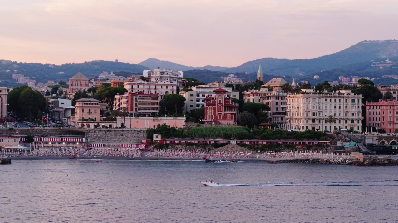 Vista aerea della costa di Genova con barche che passano vicino a edifici storici colorati al tramonto.