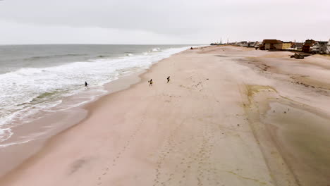 Toma-Aérea-De-Surfistas-Dejando-A-Sus-Amigos,-Corriendo-Por-La-Playa-A-Través-De-Una-Bandada-De-Gaviotas-Sosteniendo-Sus-Tablas-De-Surf