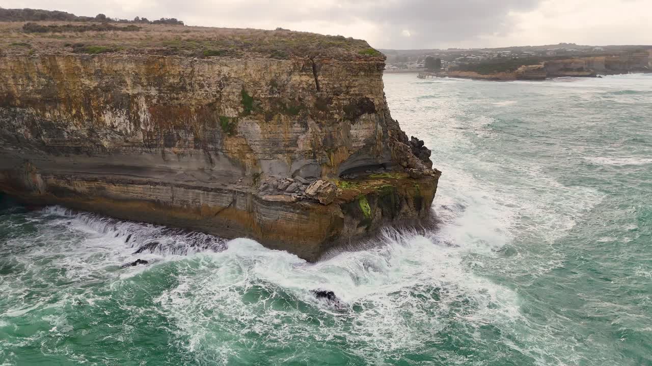 Aerial view of rugged cliffs and crashing waves at Port Campbell, Australia. Overcast skies create a dramatic coastal scene