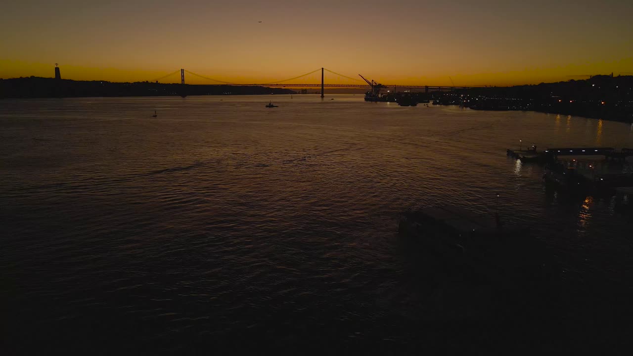 Golden hour sunset over Lisbon with calm boats on the Tagus River as the Ponte 25 de Abril appears in silhouette, backlit by warm evening light and smooth water reflections