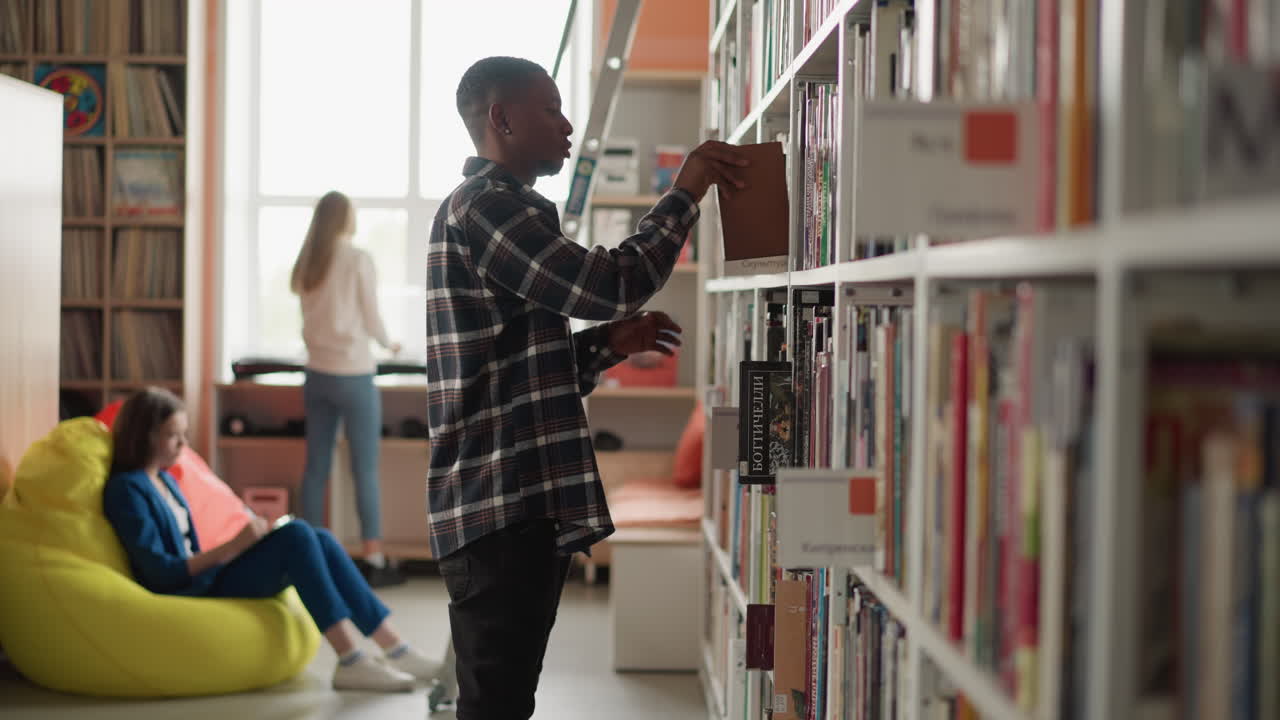 Man takes book from shelf in city public library. African American customer seeks textbook working with students in information center. Bookstore visitors