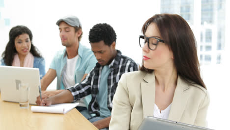 Smiling-woman-uses-tablet-while-colleagues-work-behind-her