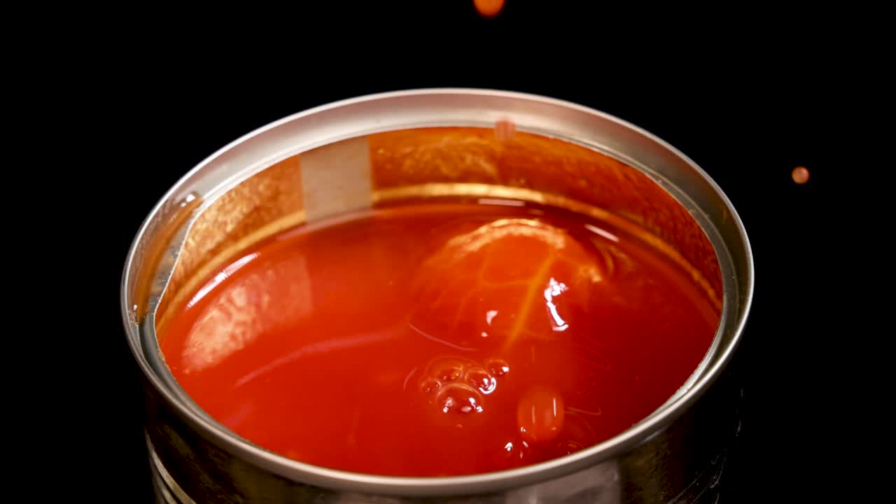 A metal fork lifts a whole peeled tomato from a can of tomato sauce, then drops it back, causing splashes under bright, even lighting with a static camera