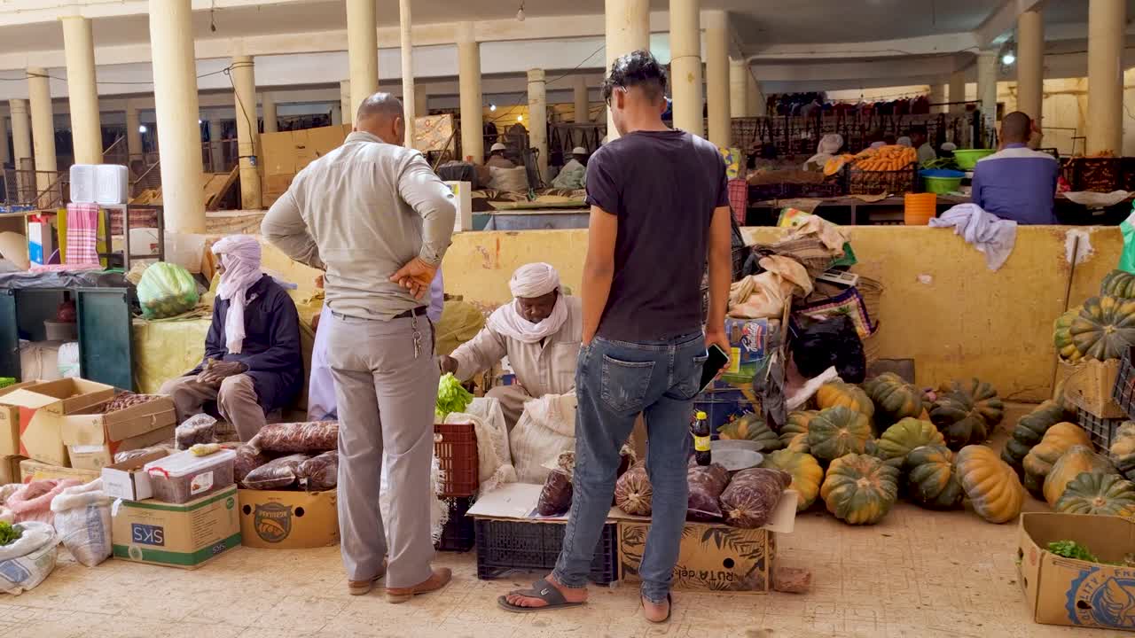 Scene at local indoor market. Man in traditional Tuareg clothing packing vegetables for customers. Filmed on sunny day in partly covered bazaar of Timimoun oasis town in the Sahara desert of Algeria