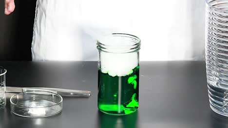 observe the reaction of a green liquid emitting vapor in a glass jar on a lab table.