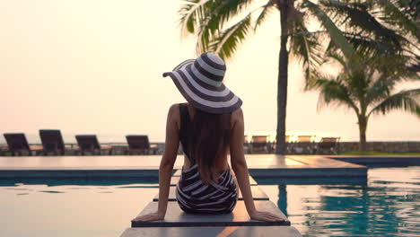 Mujer-Sexy-Sentada-En-El-Borde-De-La-Piscina-En-Un-Hotel-Exótico-En-Florida-Usando-Un-Monokini-Irregular-Y-Un-Sombrero-Rayado-Mirando-La-Playa-Al-Atardecer,-Vista-Trasera-Cámara-Lenta-De-Mano