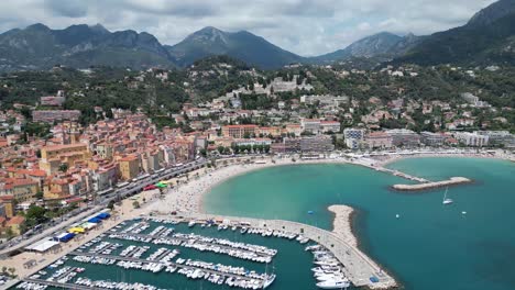 Playa-Plage-Des-Sablettes-En-Menton-Francia-Y-Casco-Antiguo-Con-Puerto-Deportivo-A-La-Izquierda,-Plano-Aéreo-A-La-Izquierda