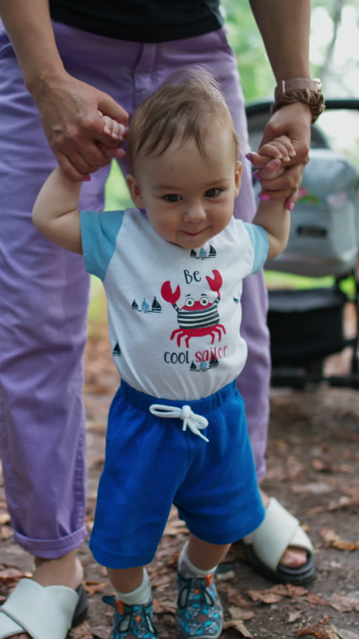 Lovely cute Caucasian boy walks supported by his mom. Little kid stepping with interest and adorable smile. Green park backdrop. Vertical video