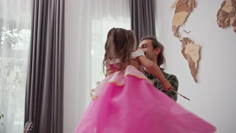 happy brunette man in a checkered green shirt picks up and spins his little daughter in a pink fairy costume in his modern home