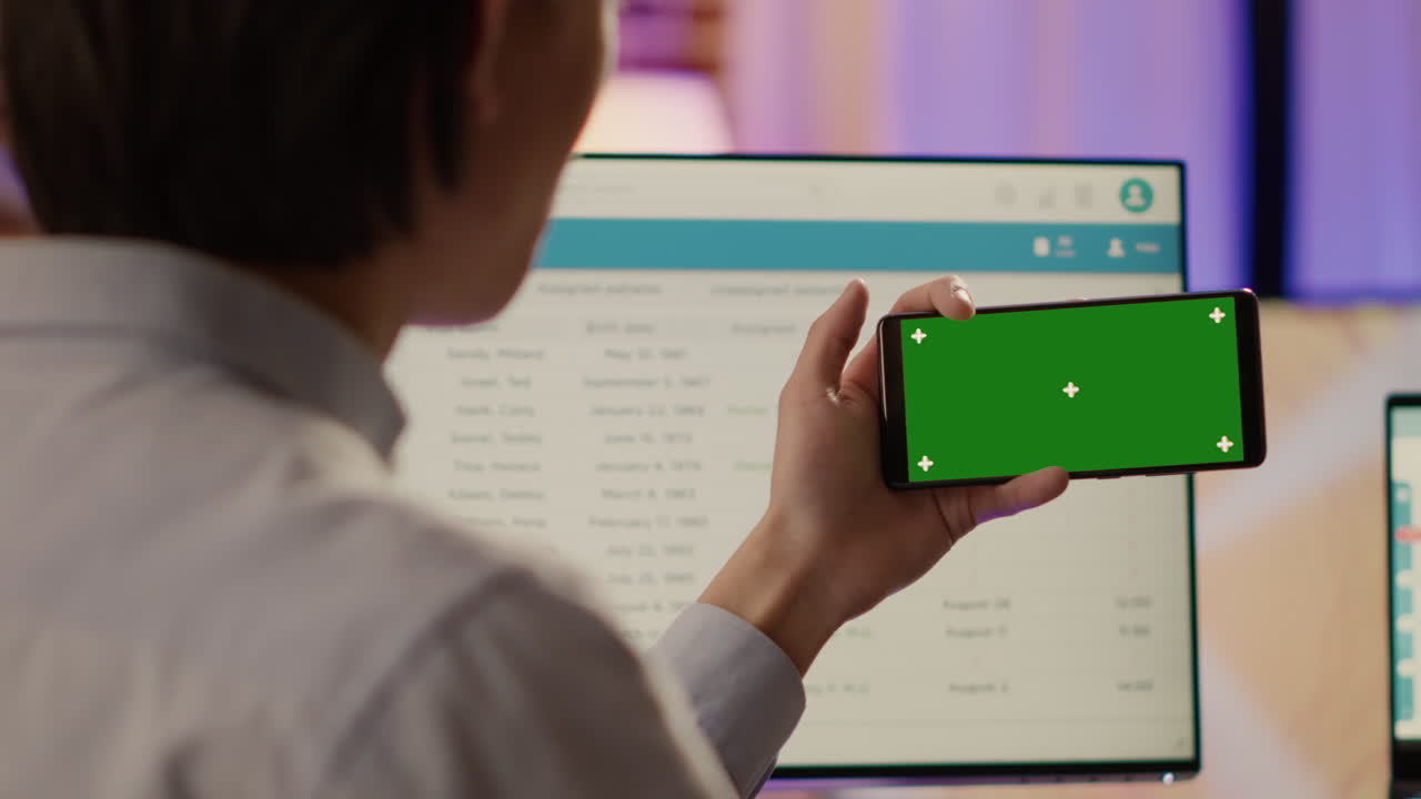 Person holding smartphone with green screen in front of computer
