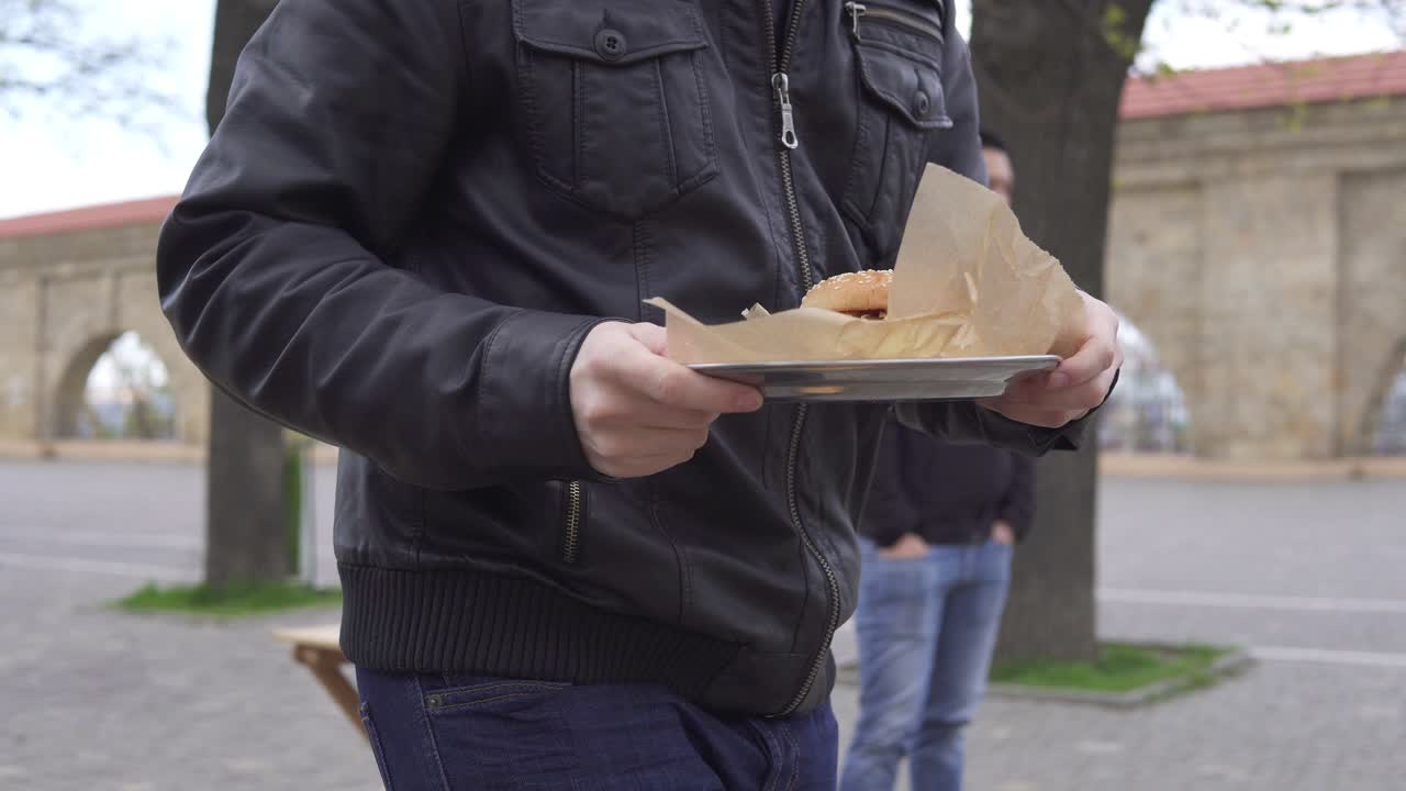 joven en un café callejero caminando a su mesa con una sabrosa hamburguesa en un gran plato, sentado. va a comer. filmado en 4k