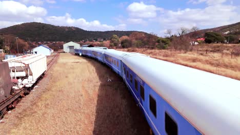 smooth rising drone shot - establishing blue botswana train , on a sunny hot day with dry grass vegetation and brown soil surrounding the railway line