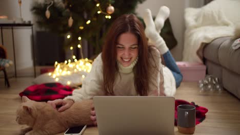feliz chica morena en un suéter blanco yace en el suelo en un cuadrilátero rojo viendo una película en una computadora portátil y acariciando a su gato de color crema en una habitación acogedora decorada en estilo navideño