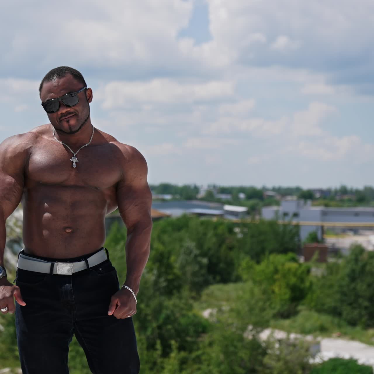 Shirtless afro american sportsman outdoors. Muscular bodybuilder with dark skin in sunglasses posing on hill on factory background