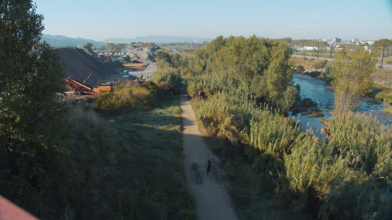 A runner runs barefoot on a dirt road, next to a river and surrounded by nature. Aerial view from a bridge