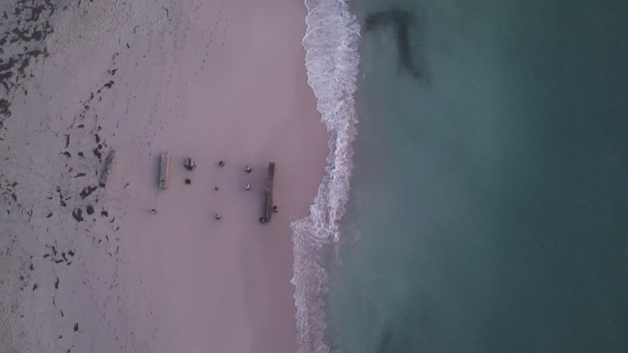 Top-down aerial footage along the beach of Jurien Bay on a winter evening. The waves crash onto the beach, dividing the scene cleanly into two halves comprising of the ocean and the sand