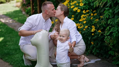 couple with a cute little baby sit squatted and kissing. parents show a number one shape balloon to their kid.