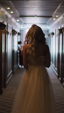 graceful woman bride in long white wedding dress with loose curly hair walks along semidark hotel hallway backside view slow motion