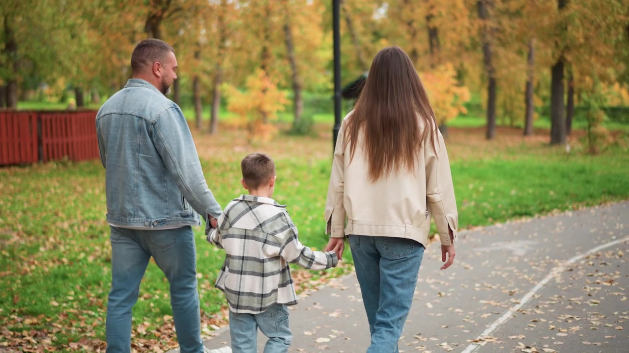 Famiglia di tre persone che cammina insieme nel parco tenendosi per mano mentre passeggiano lungo un sentiero circondati dal fogliame autunnale. La scena presenta erba verde con foglie cadute e alberi che mostrano i colori autunnali
