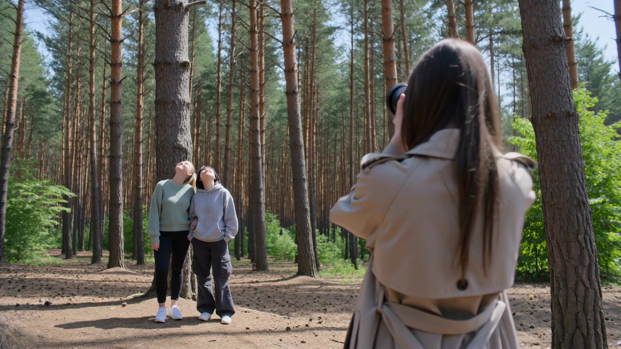 Фотограф в плаще поднимает зеркальную камеру, кадрирует и снимает друзей, позирующих в сосновом лесу для фотосессии.