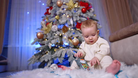 little blond baby wearing white suit sits on the floor under christmas tree. lovely child plays with a toy and pulls it to his mouth. low angle view.