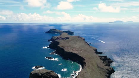 drone-view-of-an-island-rocks-with-a-distant-lighthouse-on-a-sunny-day-with-clouds-in-the-background