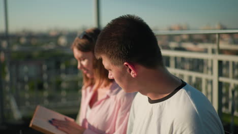 close-up of young woman showing a book to her male friend seated outdoors, his focus diverted to another direction, blurred background featuring greenery and urban landscape