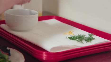 a sprig of dill and lemon zest on a white paper towel red lunch trays and a white glass bowl while a chef creates a meal in the forground