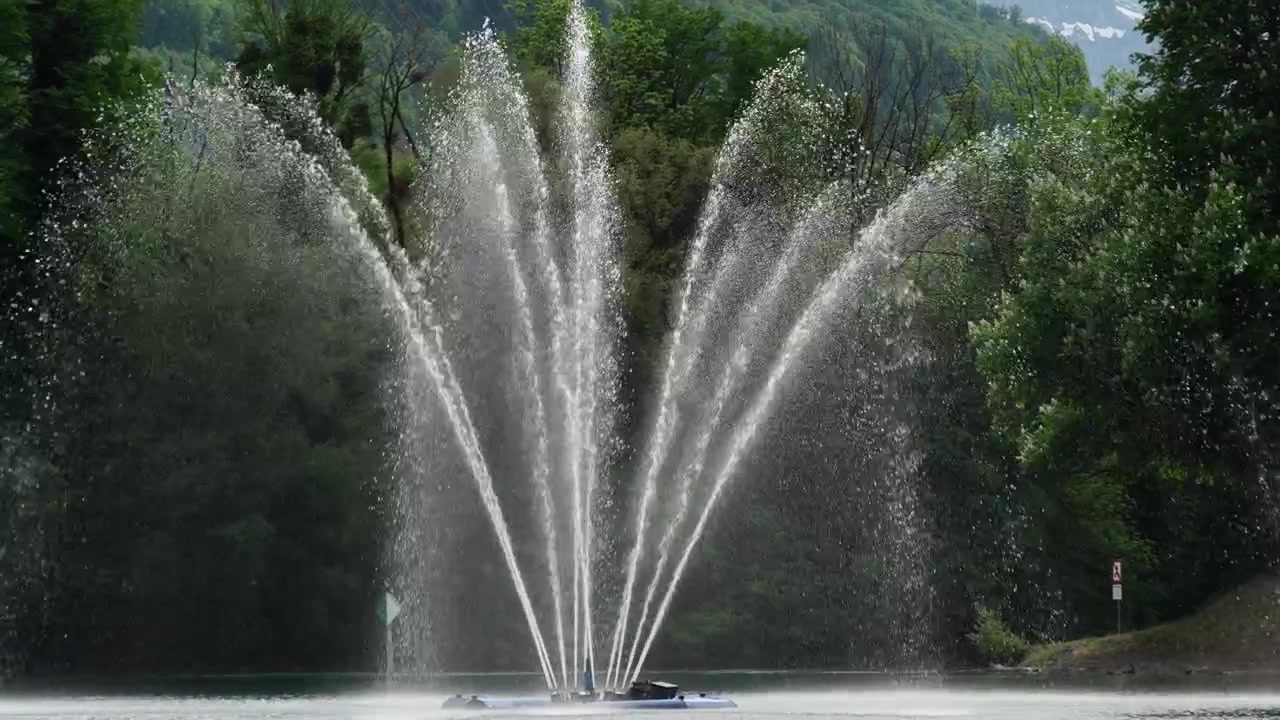 Høy buet fontene spruter vann på Walensee-sjøen, mot en skogkledd åsside i Weesen, Sveits.
