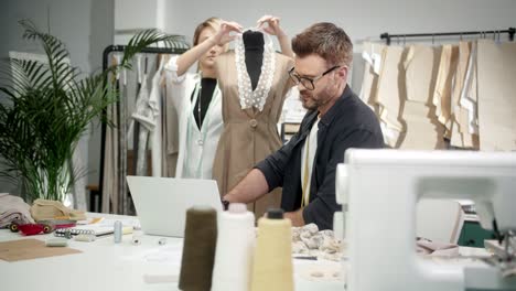 man tailor in glasses sitting at the table with a sewing machine using laptop and talking with woman colleague