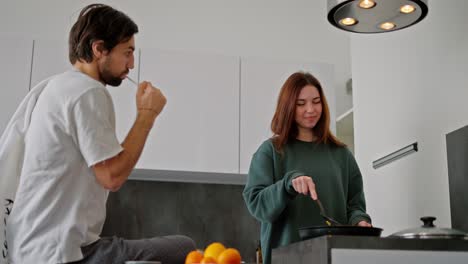 un hombre moreno con paja y en una camiseta blanca se cepilla los dientes mientras su novia morena en un suéter verde hace el desayuno en la cocina en un apartamento moderno