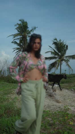 blurred woman stands on tropical beach with palm trees and a black goat in background