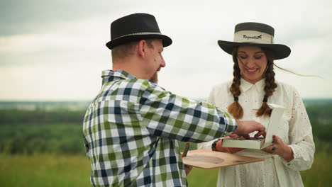 a painter wearing a black hat and plaid shirt stands in a grassy field, receiving a drawing pencil from his smiling partner. she wears a white dress and hands him the pencil with warmth and affection