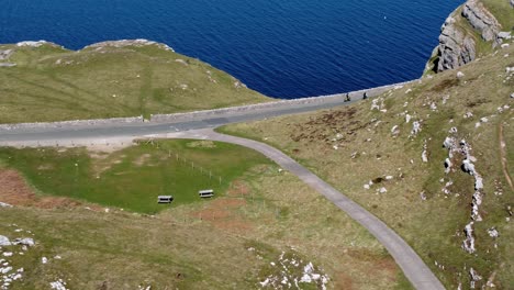 Turista-Montando-En-Bicicleta-De-Montaña-A-Lo-Largo-De-La-Pintoresca-Carretera-Rural-De-Montaña-Verde-Con-Vistas-Al-Hermoso-Mar-Azul-De-Irlanda-Panorámica-Aérea-A-La-Derecha