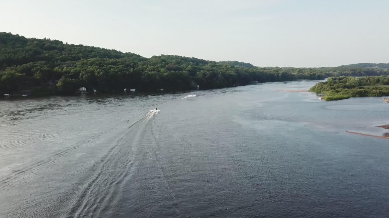 Two speedboats cut through a broad river, leaving trails in the calm water. Flanked by lush green hills, this aerial shot captures motion, recreation, and natural beauty on a sunny day.