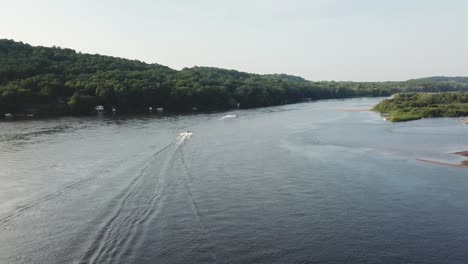 two speedboats cut through a broad river, leaving trails in the calm water. flanked by lush green hills, this aerial shot captures motion, recreation, and natural beauty on a sunny day.