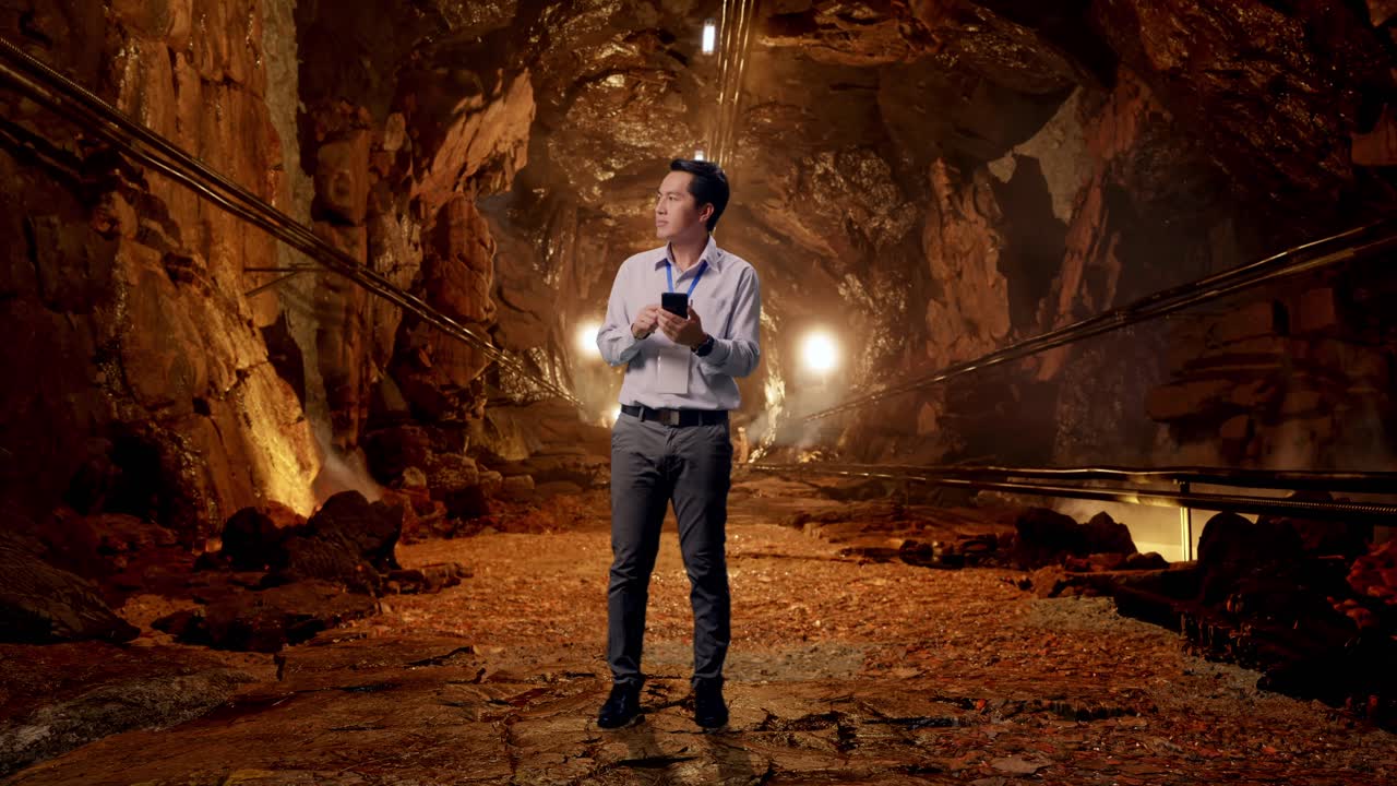 Full Body Of Asian Male Professional Worker With His Smartphone In Underground Mine Tunnel, He Observes By Looking Around Before He Come To Concentrating With His Smartphone