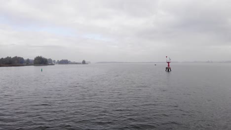 Drohne-Fliegt-über-Einen-See-Zu-Einem-Roten-Leuchtfeuer,-Das-An-Einem-Bewölkten-Tag-In-Den-Niederlanden-Vor-Seichtem-Wasser-Warnt