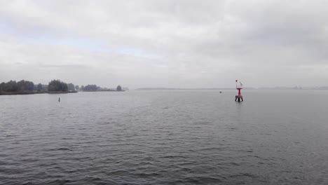 Drone-Volando-Sobre-Un-Lago-A-Un-Faro-Rojo-De-Advertencia-De-Luz-Para-Aguas-Poco-Profundas-En-Un-Día-Nublado-En-Los-Países-Bajos