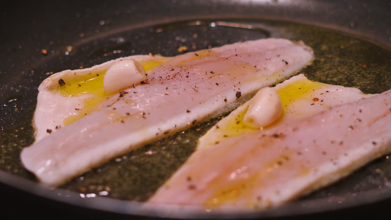 Roterende visning af to havbarsfileter på en varm pande, der steger med skindsiden nedad i boblende olivenolie. Hvid fisk tilberedt med hvidløg, salt, peber. Sund, omega-3-rig, proteinrig mad. 4K