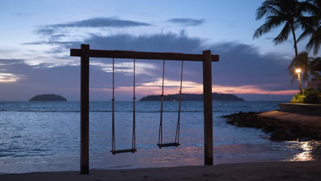 Un-Par-De-Columpios-De-Cuerda-Vacíos-En-La-Playa-De-La-Puesta-De-Sol-Junto-Al-Mar-En-Una-Isla-Tropical,-Bungalow-Con-Farolas-Y-Palmeras---Impresionante-Cielo-Dramático-En-El-Fondo