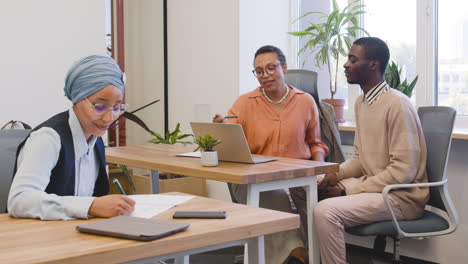 Muslim-Businesswoman-Sitting-At-A-Table-Taking-Notes-While-An-Businesswoman-Talks-To-The-New-Employee-Looking-At-Laptop-At-Another-Table-In-The-Office