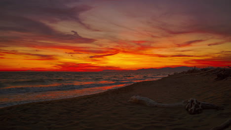 Hermoso-Bucle-De-Video-De-4k-Uhd-Cinemagraph-De-La-Puesta-De-Sol-Vista-Desde-Una-Romántica-Playa-De-Arena-Con-Madera-A-La-Deriva-Y-Olas-En-La-Costa-Mediterránea-Italiana-Con-Nubes-Rojas-Y-Naranjas-En-El-Cielo