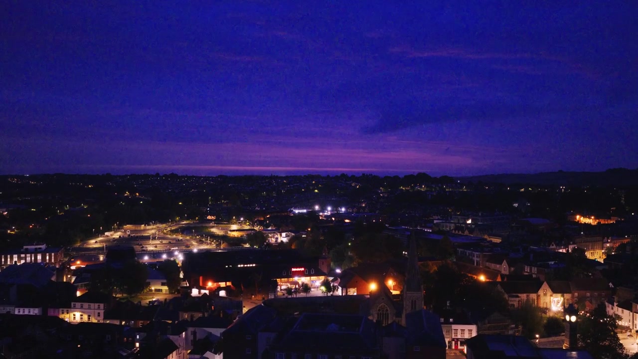 Flyfoto over Salisbury i den blå timen som viser opplyste gater, bygninger og hustak under en dyp blå og lilla kveldshimmel