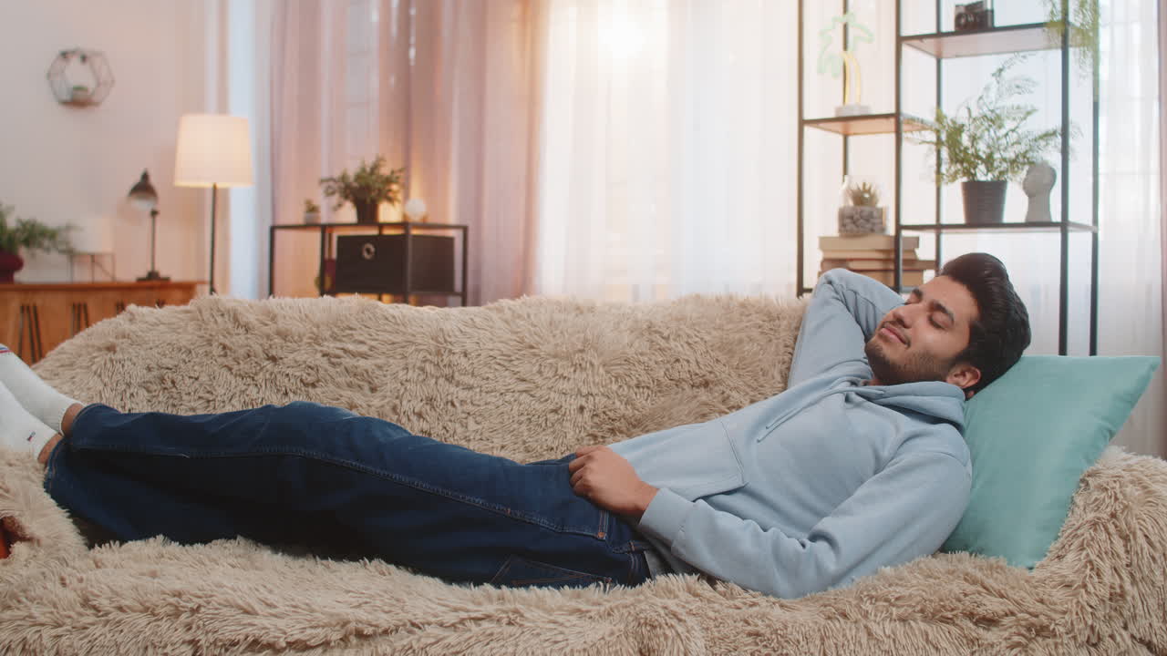 Tired indian man lying on comfortable couch taking rest and daytime nap in living room at home
